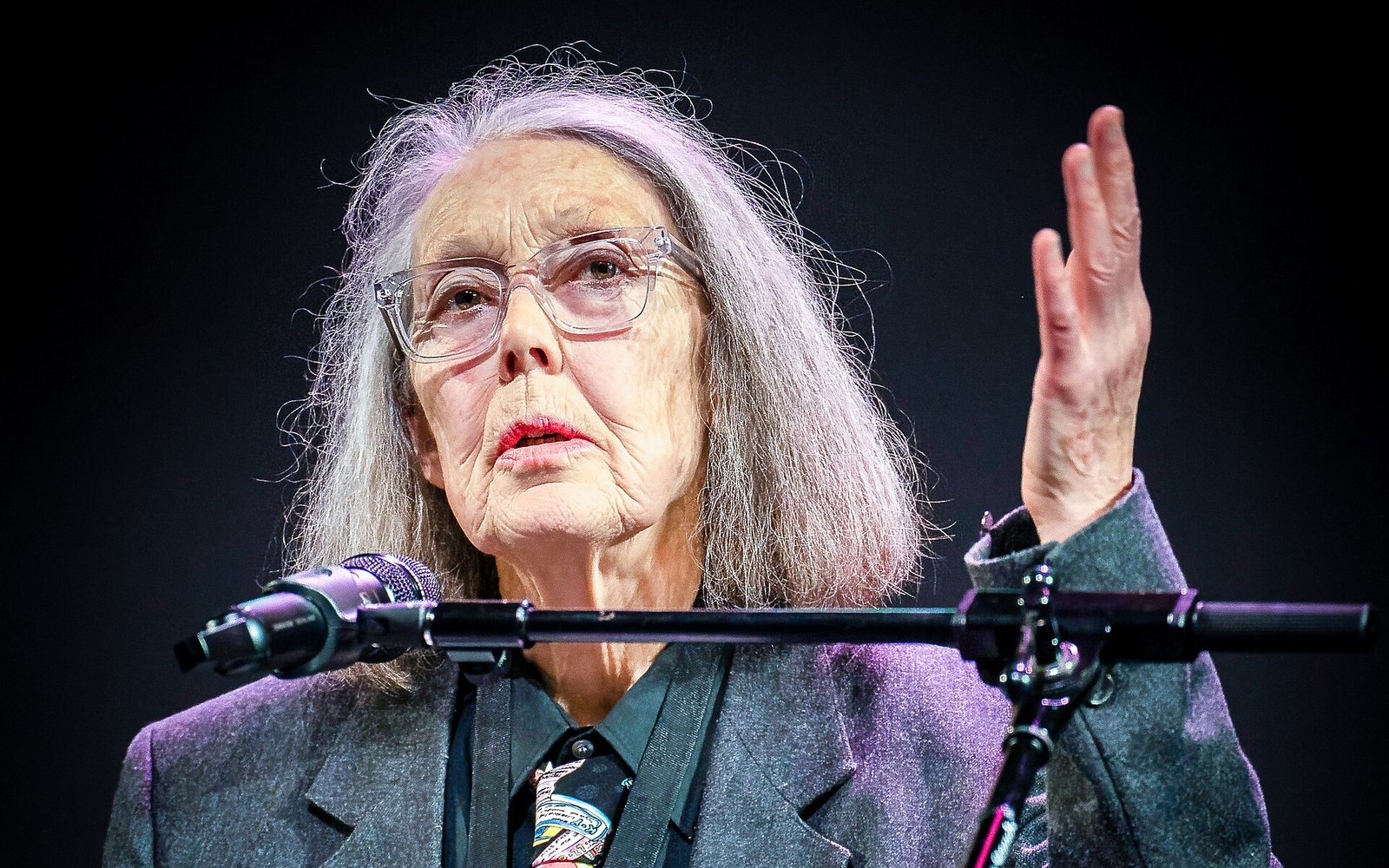 Anne Carson, med grått hår och glasögon, talar i en mikrofon från en scen och gestikulerar med handen