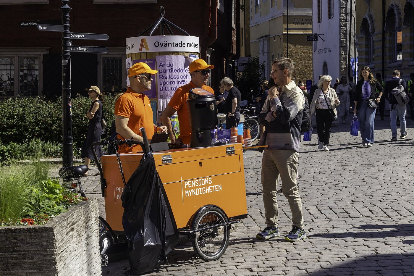 Två medarbetare från Pensionsmyndigheten i orange kepsar och tröjor står vid en orange lådcykel med myndighetens logga och pratar med en man på en kullerstensgata i Visby under Almedalsveckan